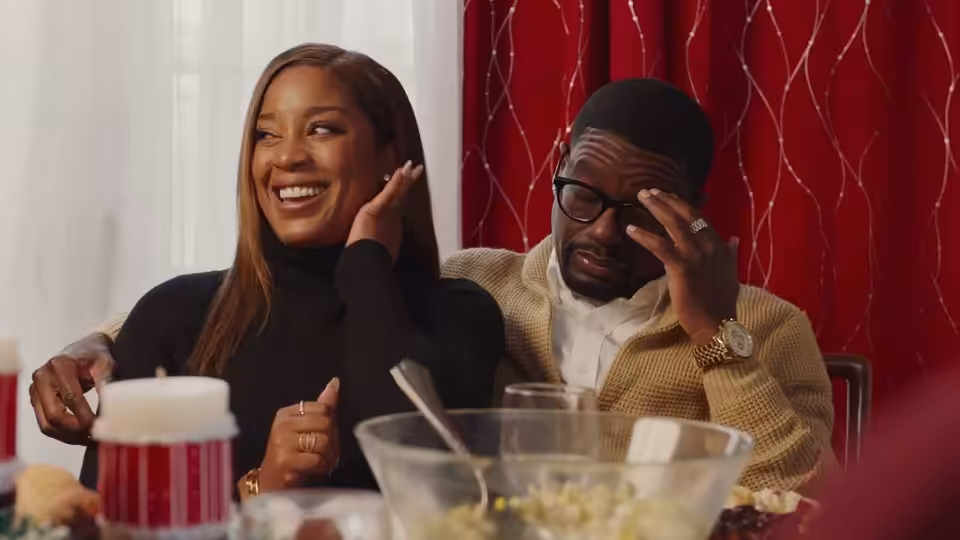 A woman laughs beside a bespectacled man at a dinner table as he rubs his eye