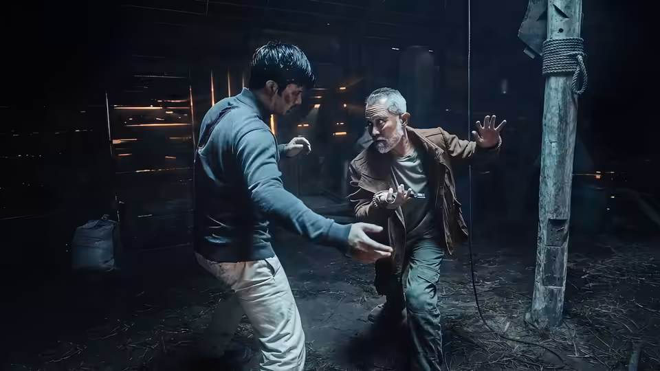 A younger man faces an older gray-haired man in a fighting stance inside a dim wooden hut