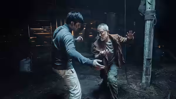 A younger man faces an older gray-haired man in a fighting stance inside a dim wooden hut