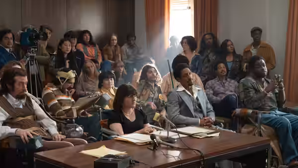 A man addresses a crowded meeting as attendees sit around a conference table