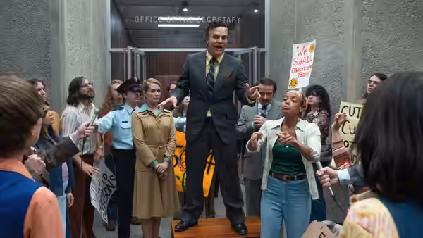 A suited man addresses disability-rights demonstrators in a government corridor