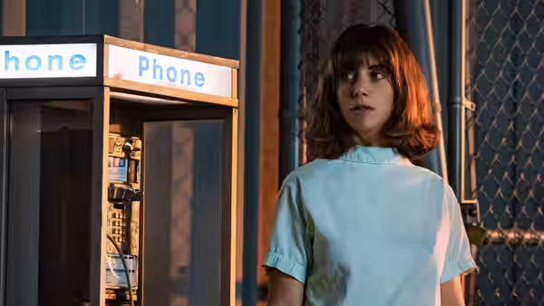 Sarah (Alison Brie) stands beside a public phone booth in a pale blue gown