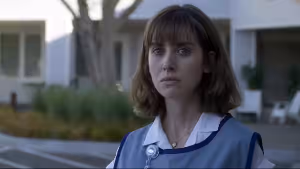 Sarah (Alison Brie) stands outside a house wearing a blue-and-white care uniform
