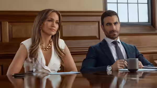 Jackie Cruz (Jennifer Lopez) and Daniel Blanchflower (Brett Goldstein) sit together at a conference table