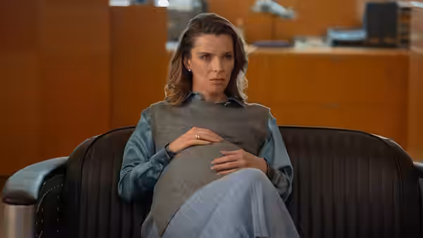 A woman sits tensely in a leather chair, clutching a cushion in an office