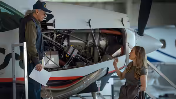 A woman gestures toward an exposed engine as an older man examines the aircraft in a hangar