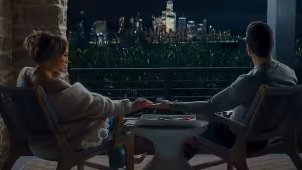 A couple sit holding hands on a terrace overlooking the city skyline at night