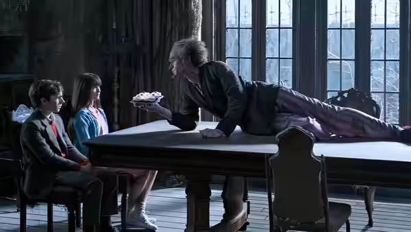 A gray-haired man leans across a table offering cake to Violet and Klaus Baudelaire