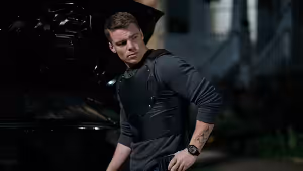 Peter Sutherland (Gabriel Basso) stands beside a car wearing a tactical vest