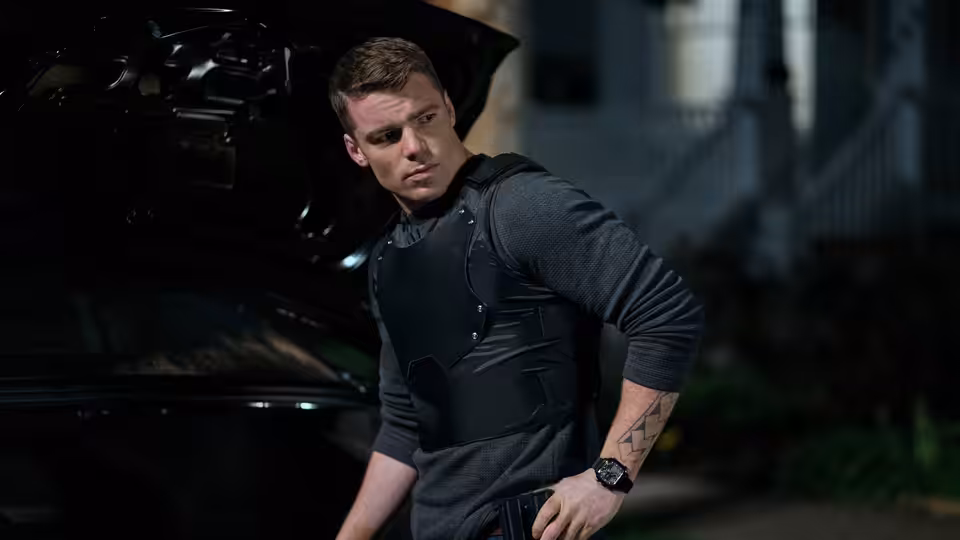 Peter Sutherland (Gabriel Basso) stands beside a car wearing a tactical vest