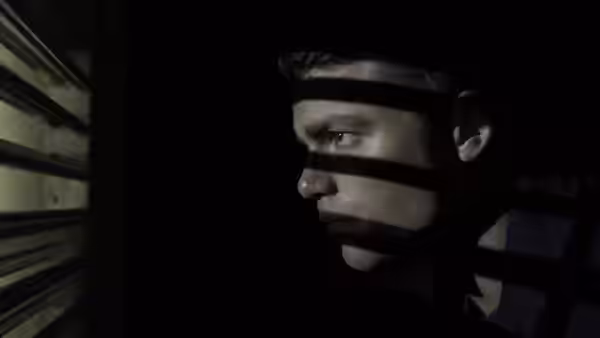 A man peers through window blinds as bands of shadow cross his face