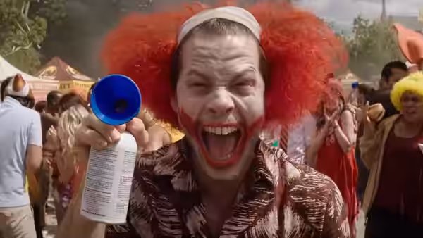 A man in a red clown wig yells through a megaphone at a crowded outdoor party