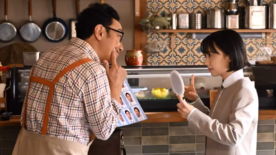 Minamida Minami (Mana Ashida) holds up a hand mirror as a man examines photographs in a kitchen