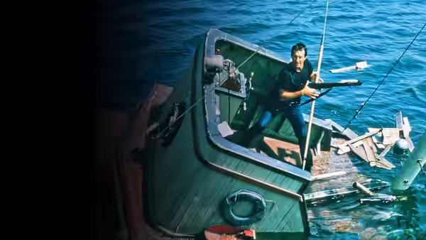 Quint aims a rifle from the deck of a green boat on the water