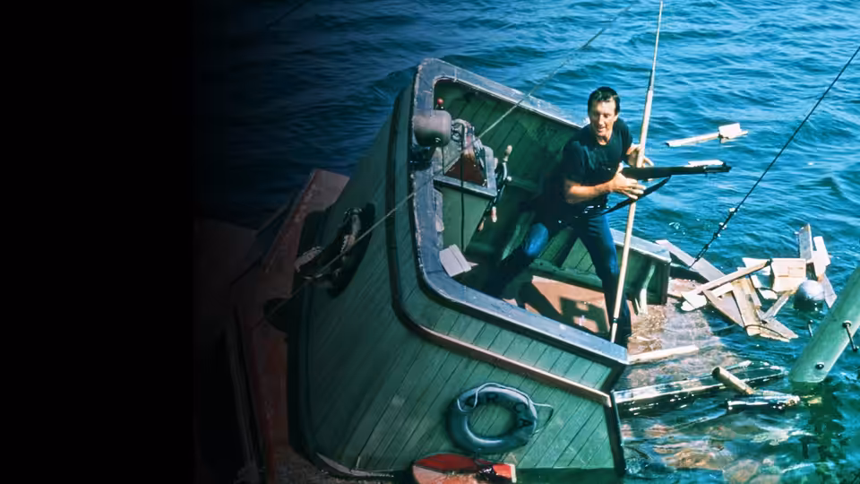 Quint aims a rifle from the deck of a green boat on the water