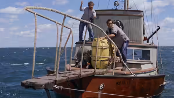 Quint waves from atop a boat while Hooper handles a yellow barrel