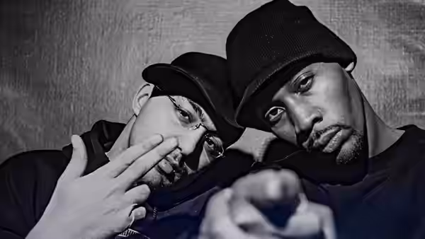 RZA in a beanie points toward the camera beside a man wearing a cap and glasses