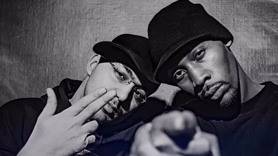 RZA in a beanie points toward the camera beside a man wearing a cap and glasses