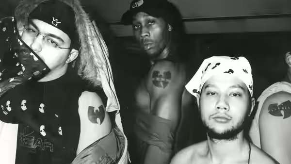RZA stands shirtless between two men indoors, one wearing a bandana and the other a head covering