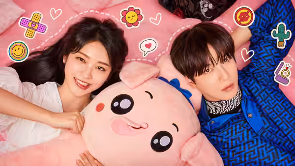A woman and a man lie beside a large pink plush toy, looking toward the camera