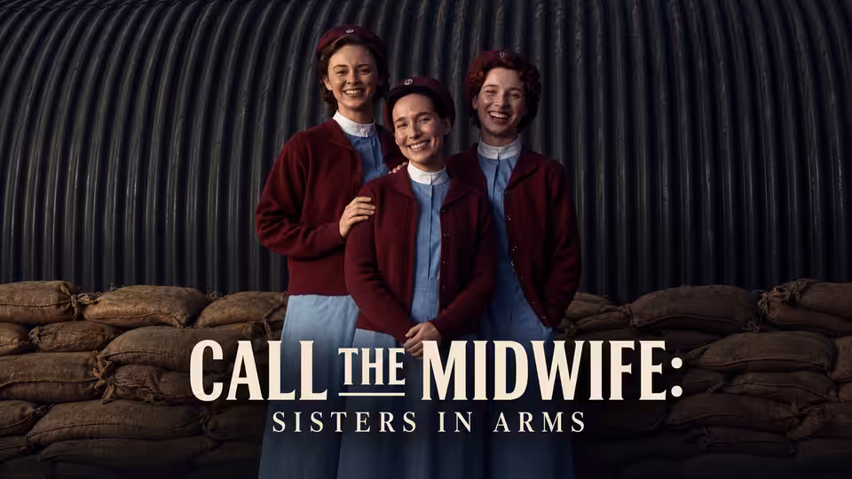 Three nurses in burgundy cardigans pose together before a corrugated metal wall and sandbags
