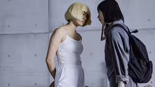 A blonde woman and a backpacked young woman face each other against a concrete wall
