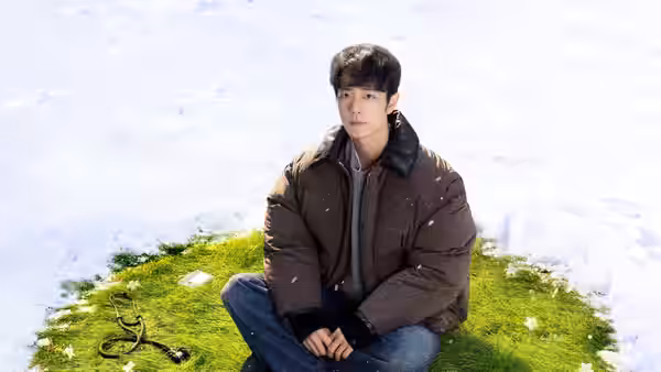 A young man sits on a patch of grass surrounded by snow