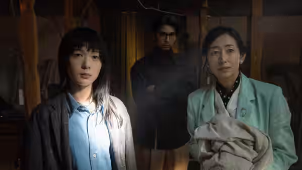 Two women and a bespectacled man stand in a wooden room, one woman holding a bundle