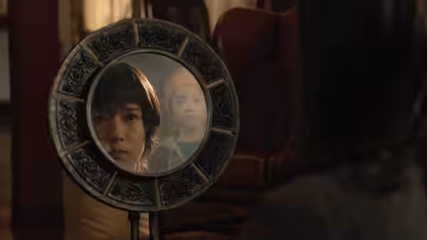A woman looks into an ornate mirror as another face appears in its reflection