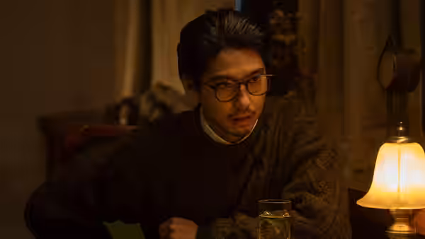 A bespectacled man sits at a table beside a glowing lamp