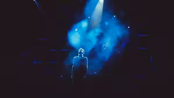 A lone performer stands beneath a spotlight amid swirling blue stage smoke