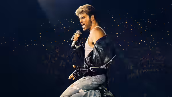 George Michael sings into a microphone onstage, wearing a leather jacket before a packed audience