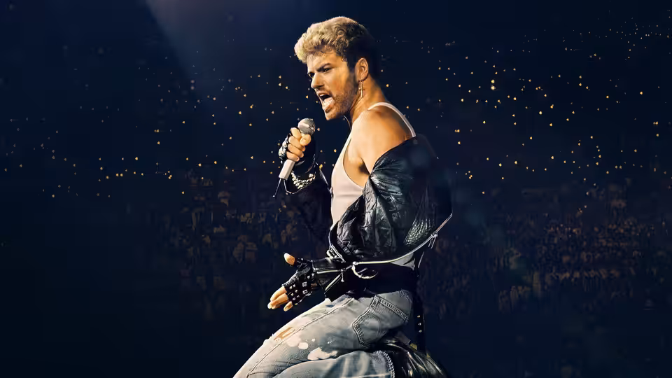 George Michael sings into a microphone onstage, wearing a leather jacket before a packed audience