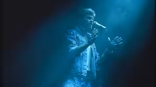 George Michael sings into a microphone, raising his hands under blue stage lights
