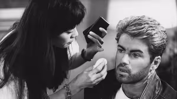 A makeup artist applies powder to George Michael's face as he looks toward the mirror