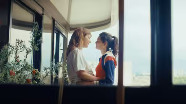 Two women stand close together beside a bright apartment window