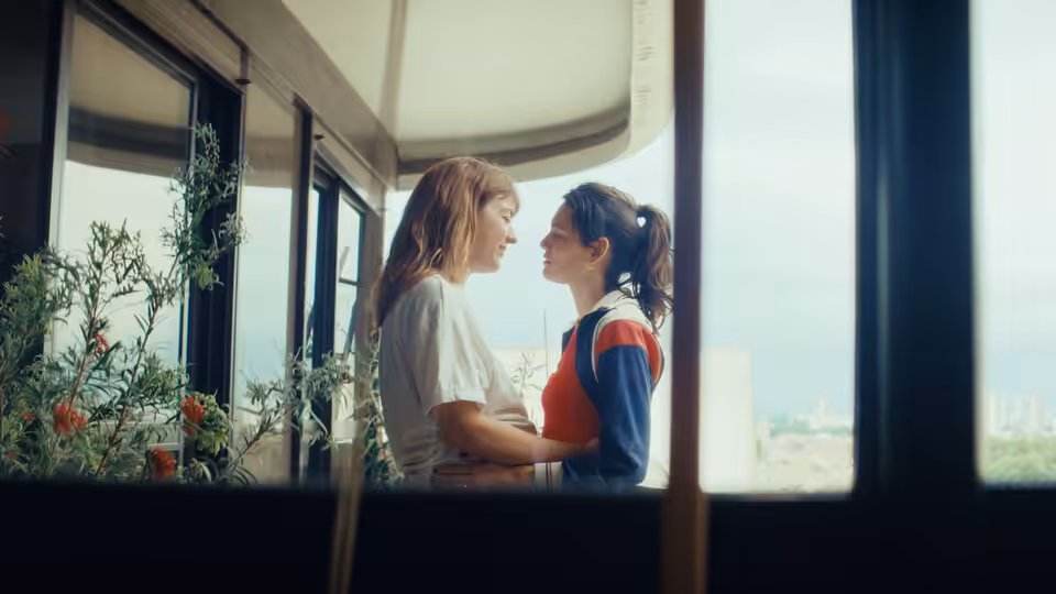 Two women stand close together beside a bright apartment window