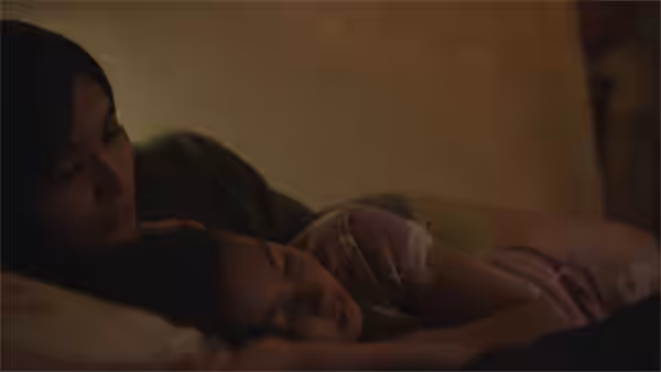 A woman lies beside a sleeping child beneath blankets in a dim room