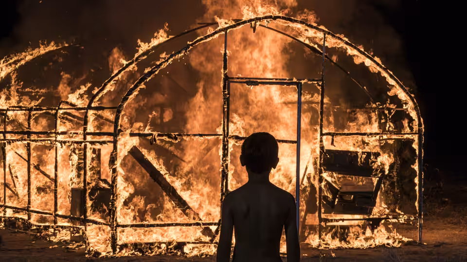 a young man stands before a large structure engulfed in flames at night