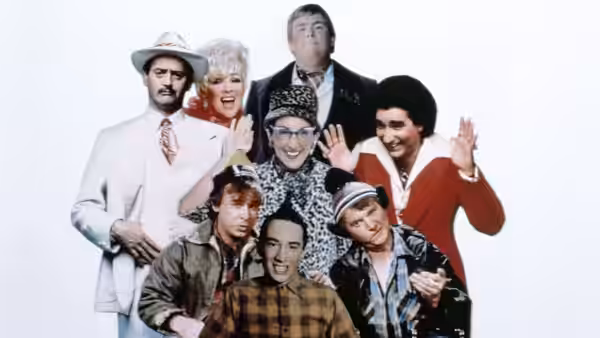 A group of performers in varied costumes pose together, several waving against a white backdrop