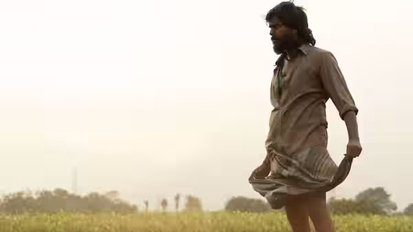 A bearded man strides through a field, lifting his lungi above the grass