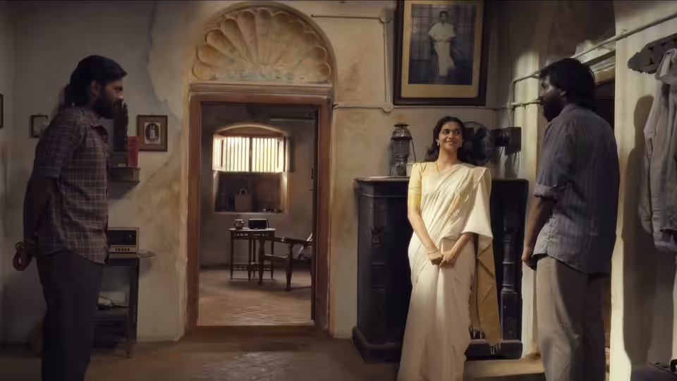 A woman in a white sari stands between two men in a worn interior