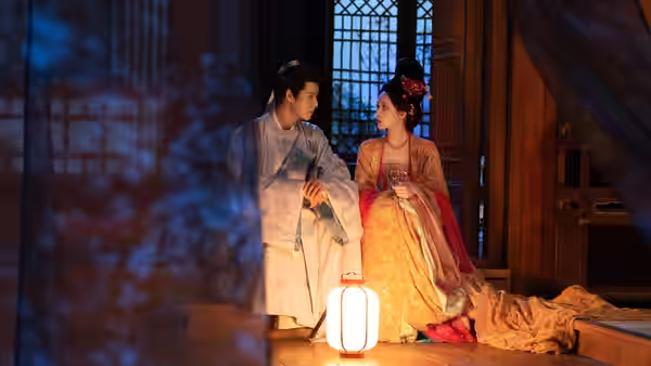 A young man and woman in traditional robes sit together beside a glowing lantern