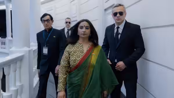 A woman in a green sari walks through a white corridor with two suited men behind her