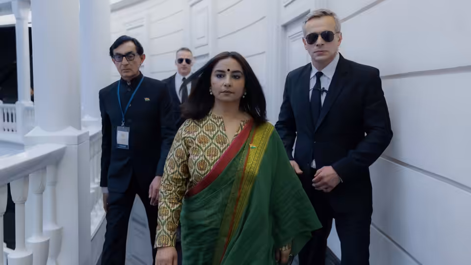 A woman in a green sari walks through a white corridor with two suited men behind her