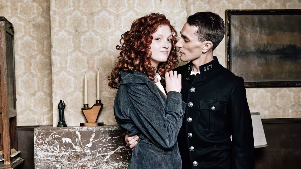 A red-haired woman and a uniformed man stand close beside a marble fireplace