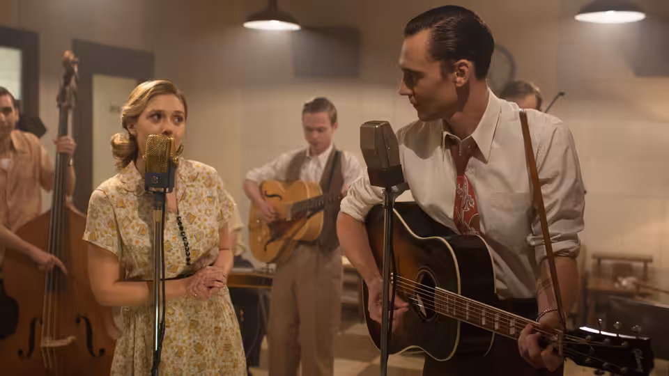 Hank Williams (Tom Hiddleston) plays guitar beside Audrey Mae Williams (Elizabeth Olsen) at a recording microphone