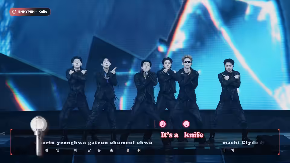 Six performers in black outfits dance onstage before a blue LED screen
