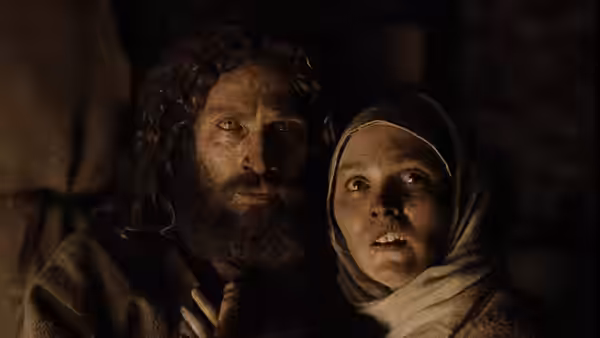 A bearded man and a headscarf-wearing woman huddle together in a dim room