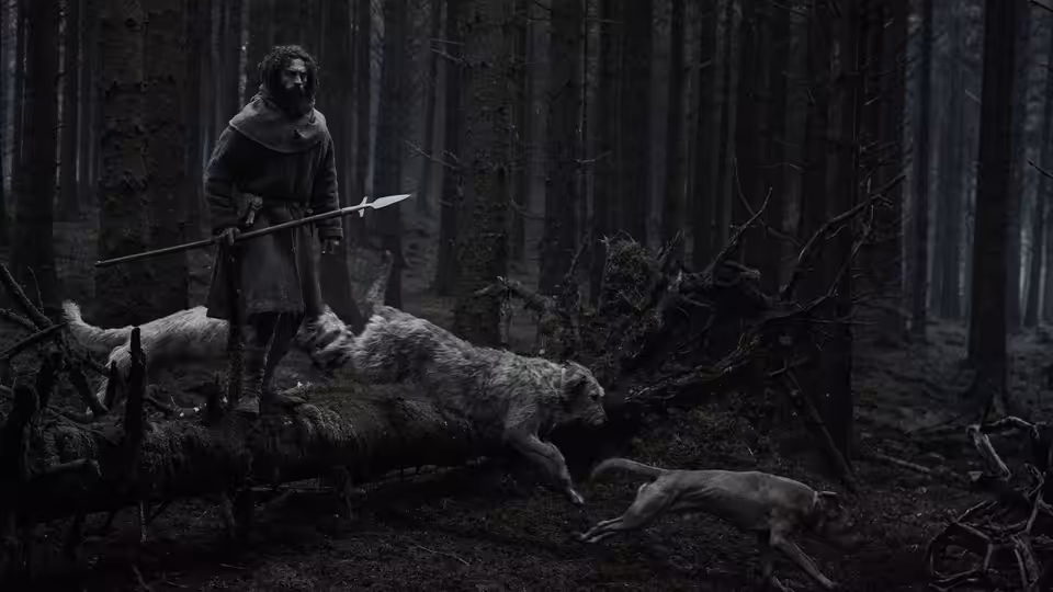A cloaked man stands among fallen animals in a dark forest, holding a spear